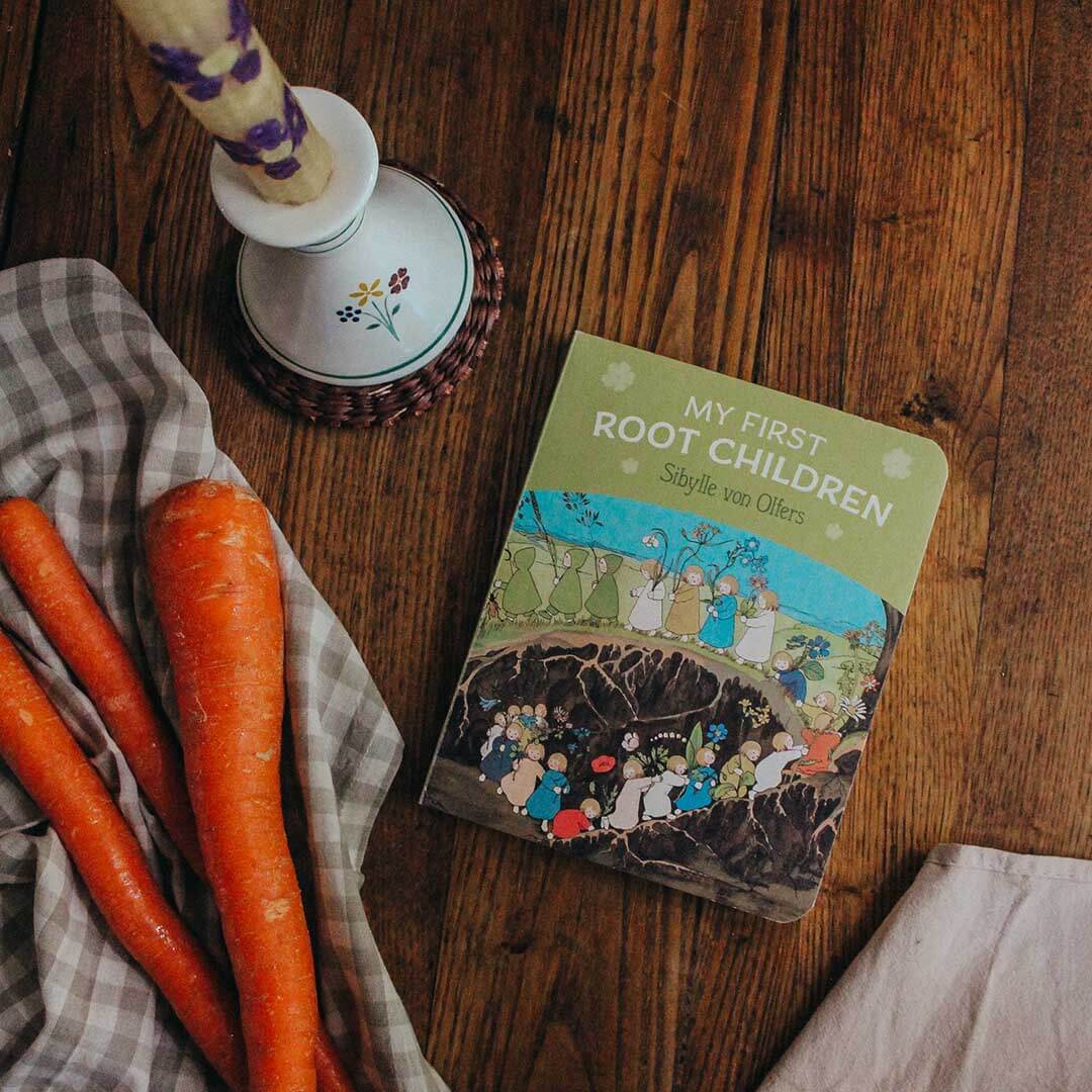 Children's book titled 'My First Root Children' on a wooden table with carrots and a checkered cloth.
