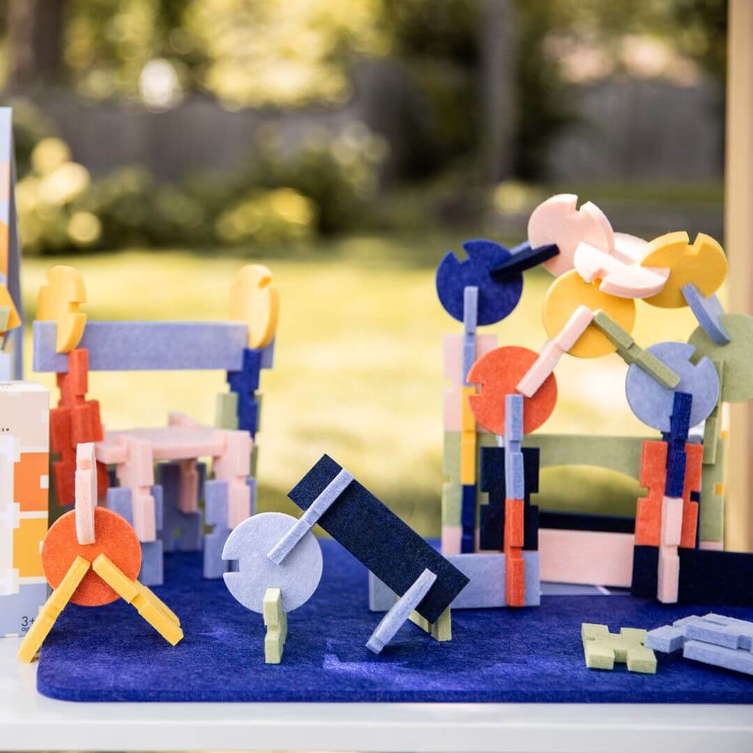 Colorful structures built with the Architectural Felt Block Set from Lowercase Toys displayed outdoors on a blue mat.