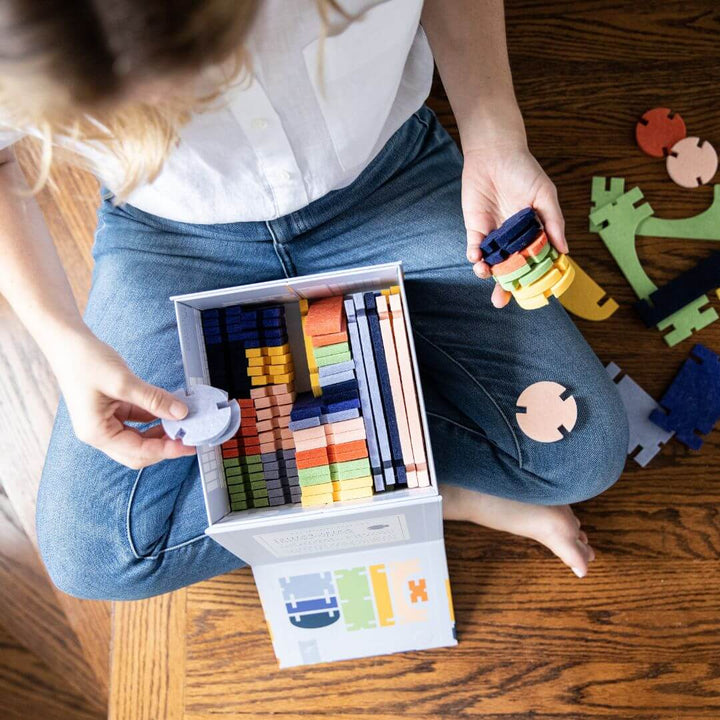 Child sitting on the floor holding felt block pieces while organizing the Felt Architectural Building Set fro Lowercase Toys inside its storage box.
