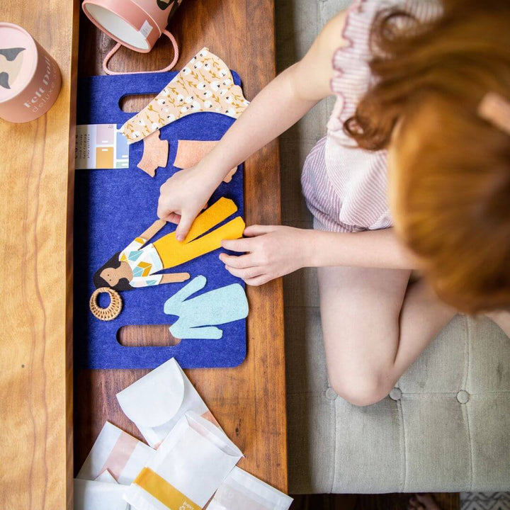 Child seated at a table, dressing felt dolls with clothing pieces from Lowercase Toys on a blue felt play mat.