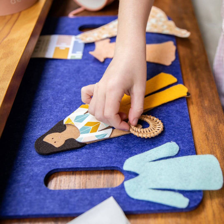 A hand shown playing with Lowercase Toys Deluxe Felt Doll Set with the clothing and doll placed on a thick blue felted mat. 