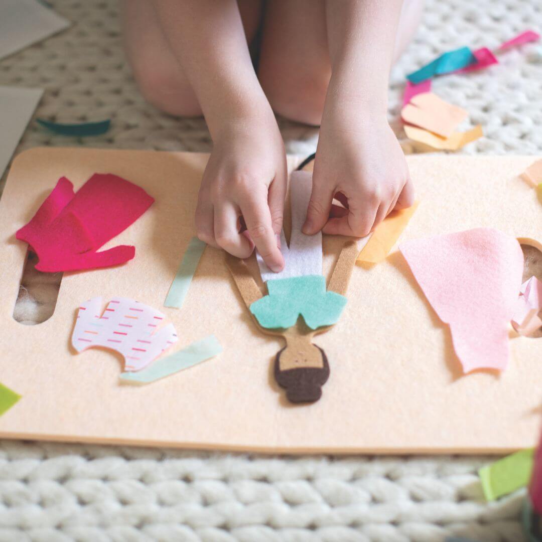 Child’s hands placing felt clothing on a felt doll from Lowercase Toys with mix-and-match outfit pieces spread out on a play mat.