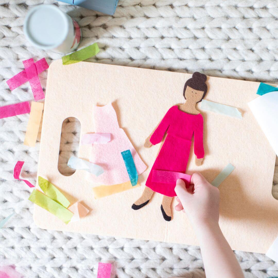 Child’s hands placing a piece of confetti and felt clothing on a felt doll from Lowercase Toys with mix-and-match outfit pieces spread out on a play mat.