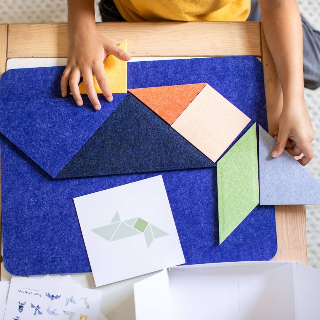 Child arranging colorful Tangram felt pieces from Lowercase Toys on a blue play mat, with a pattern guide card nearby.