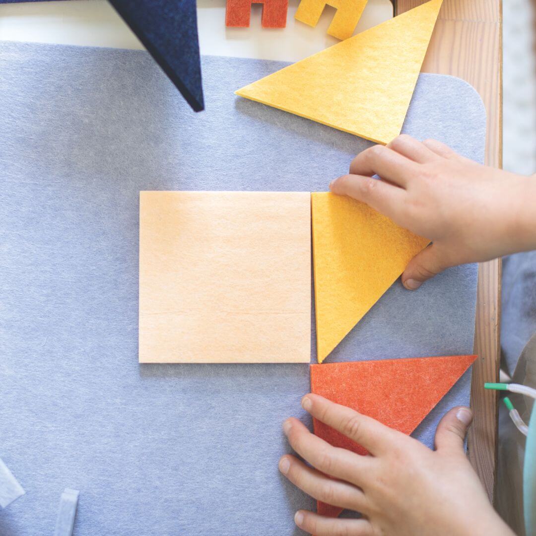 Child holding felt Tangram puzzle pieces on a pale blue felt board from Lowercase Toys while seated at a table, ready to play.