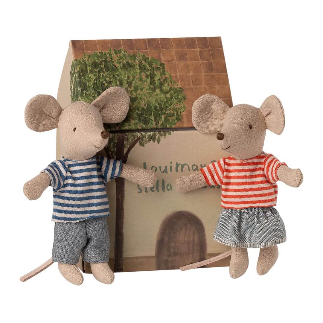 Two plush Maileg mouse toys, one in a striped shirt and shorts, the other in a dress, standing in front of a wooden house with a tree.