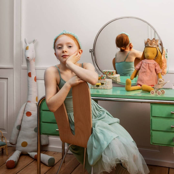 Young girl in a dress sitting at a vanity mirror with a stuffed toy and dresser in the background.