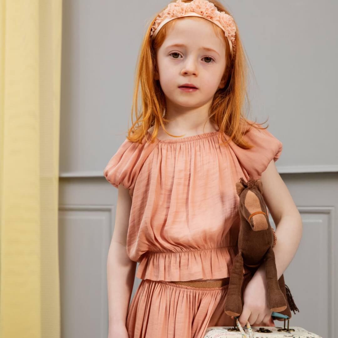 Young girl in a peach dress and headband standing against a neutral background