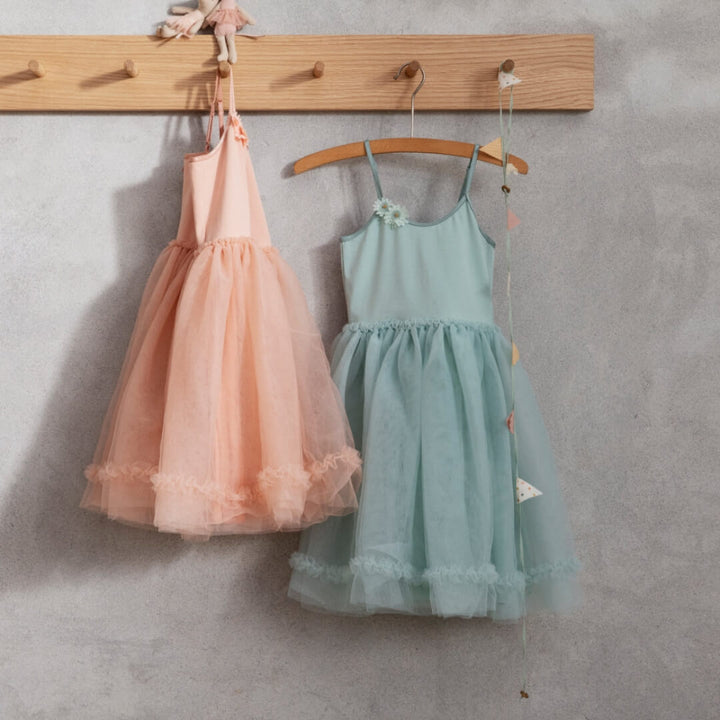 Two children's dresses, one peach and one mint green, hanging on a wooden rack against a gray wall.