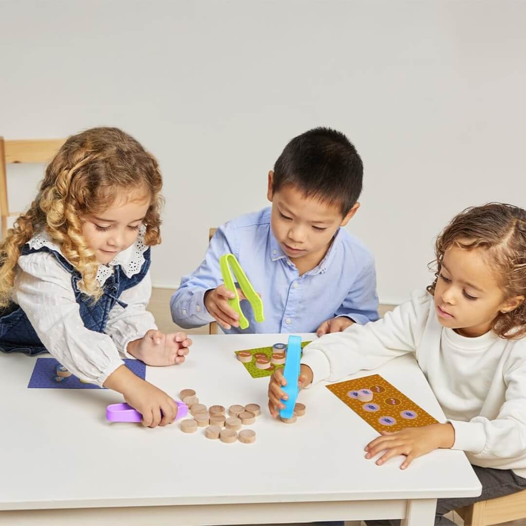 Three children sitting at a table playing the Catch the Bug! game from Miniland, using colorful tweezers to pick up wooden bugs and match them to their game boards.