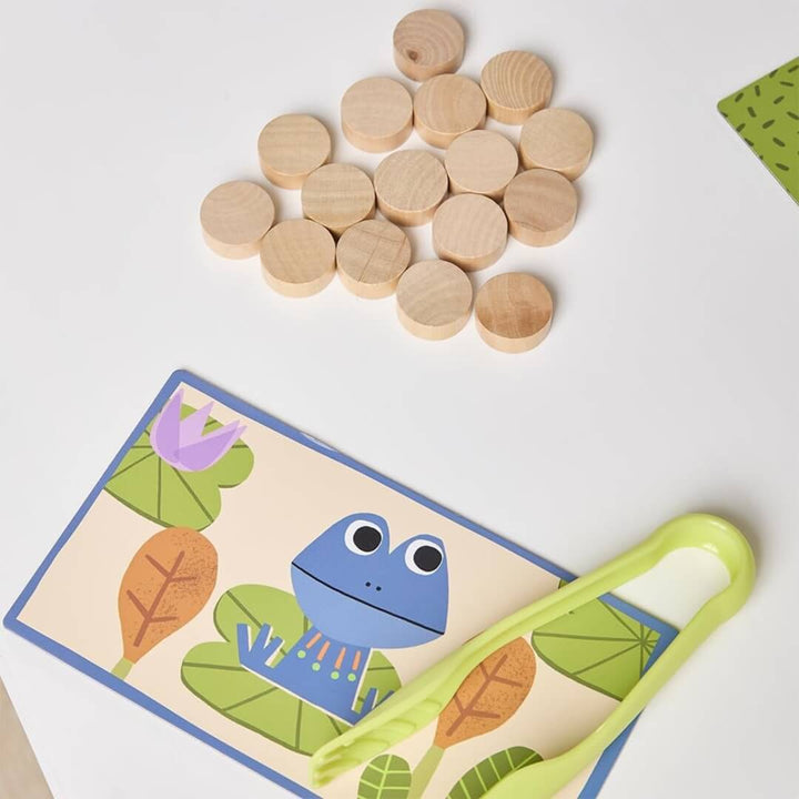 A close up of one of the board games with a frog on it, green tweezers, and a bunch of wooden discs on a white background.