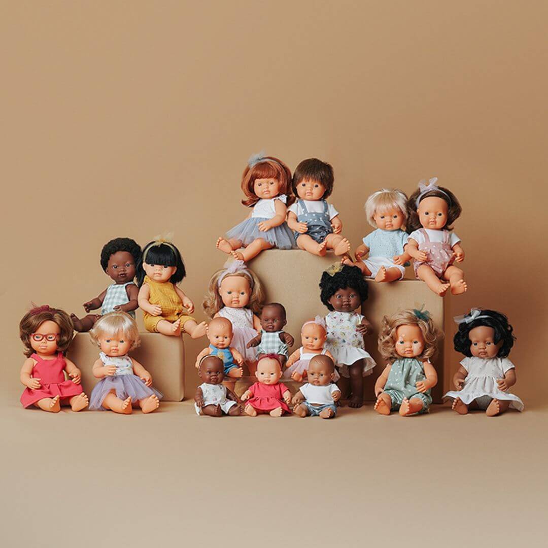 A collection of Miniland Dolls sitting on various platforms and showing the different sizes, diversity and range of clothing, skin and hair color and differences. 