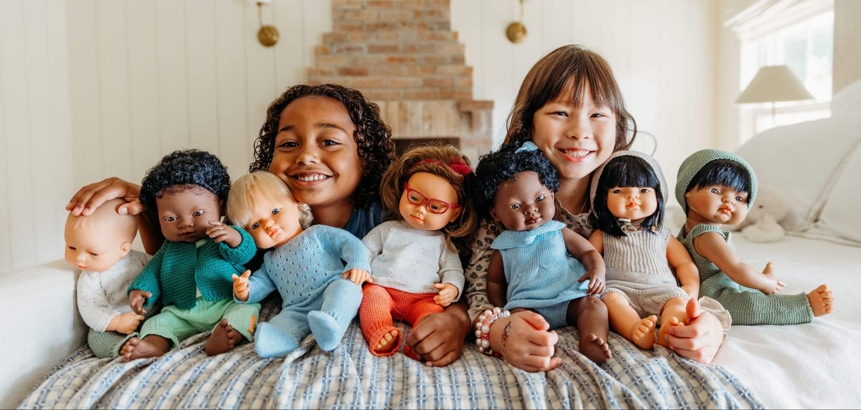 Two girls sitting on a bed with a collection of diverse dolls.