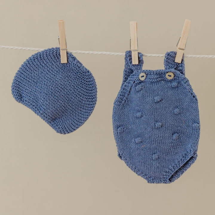 Blue knitted baby hat and romper on a clothesline against a beige background