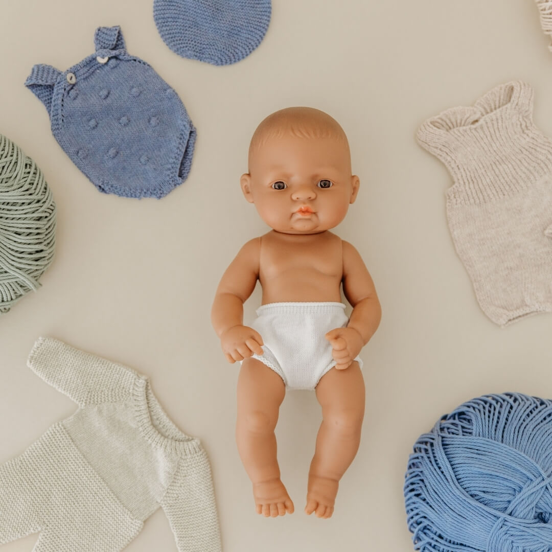Miniland doll on a cream background, surrounded by knit baby doll clothes.