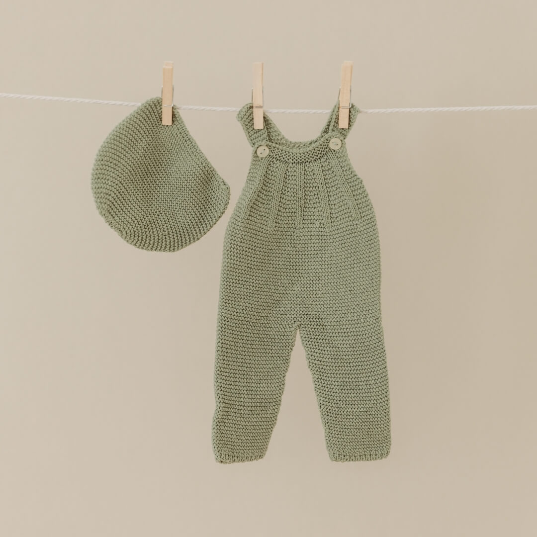 Knitted green baby overalls and hat hanging on a line against a beige background