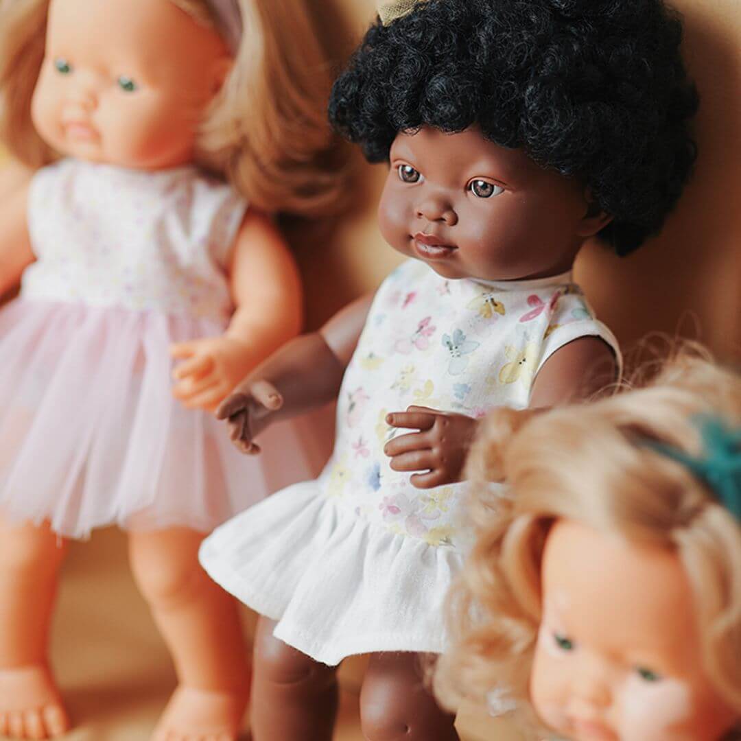 A close up of the Realistic Montessori Black Girl Doll leaning against an orange wall with two other dolls on either side. 