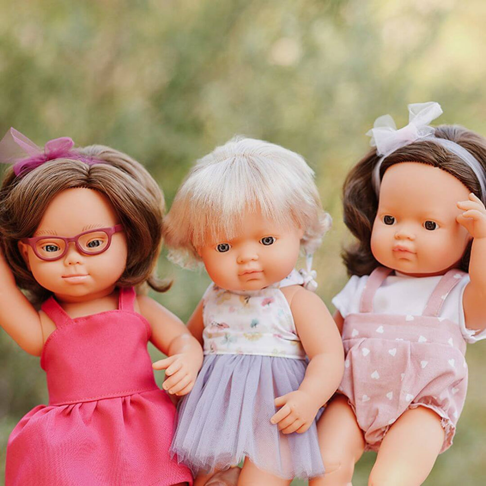 Three Miniland realistic Montessori dolls standing side by side outdoors, each wearing colorful outfits with bows and tulle, shown in front of a blurred green background of trees.