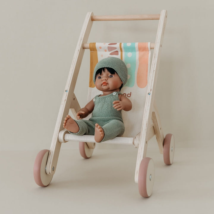 Doll in a green outfit sitting in a wooden doll stroller by Miniland, made in Spain from sustainable wood with a colorful fabric seat against white background.