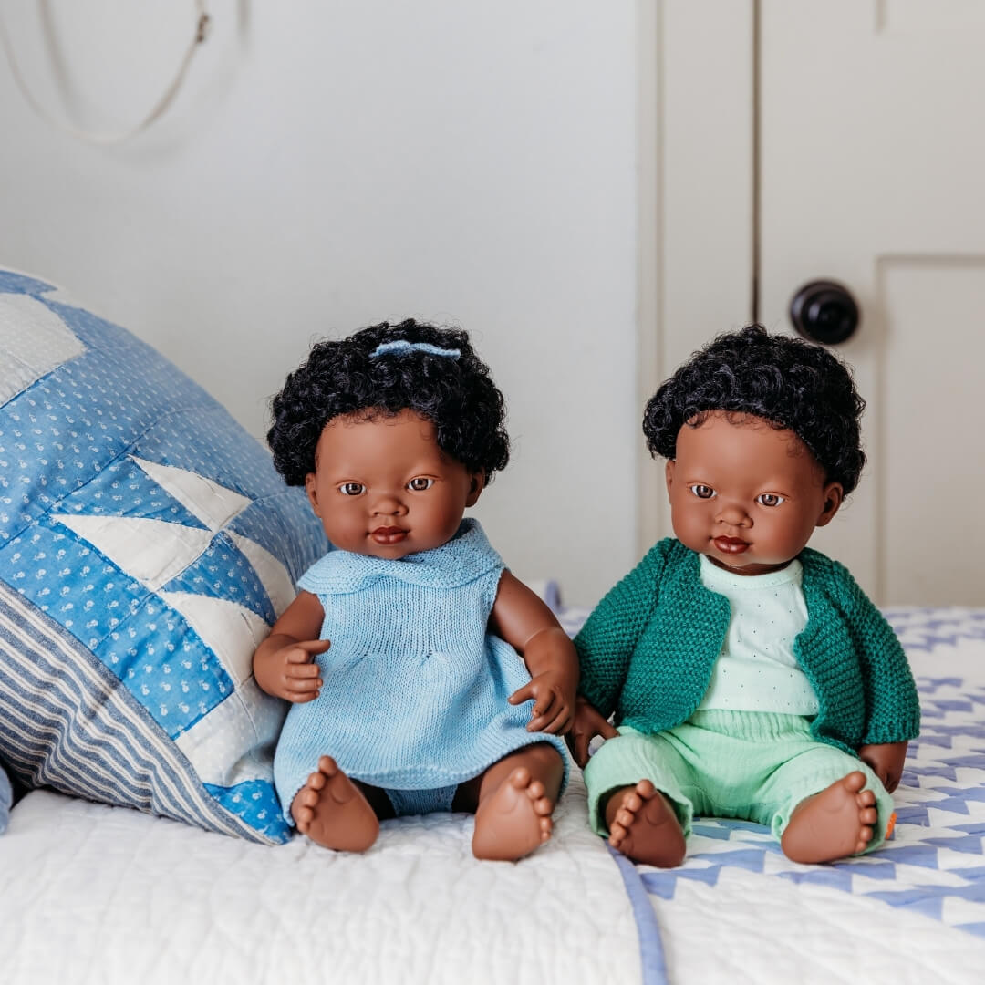Two black Miniland dolls in knit clothing sitting on a bed with a blue and white quilt.