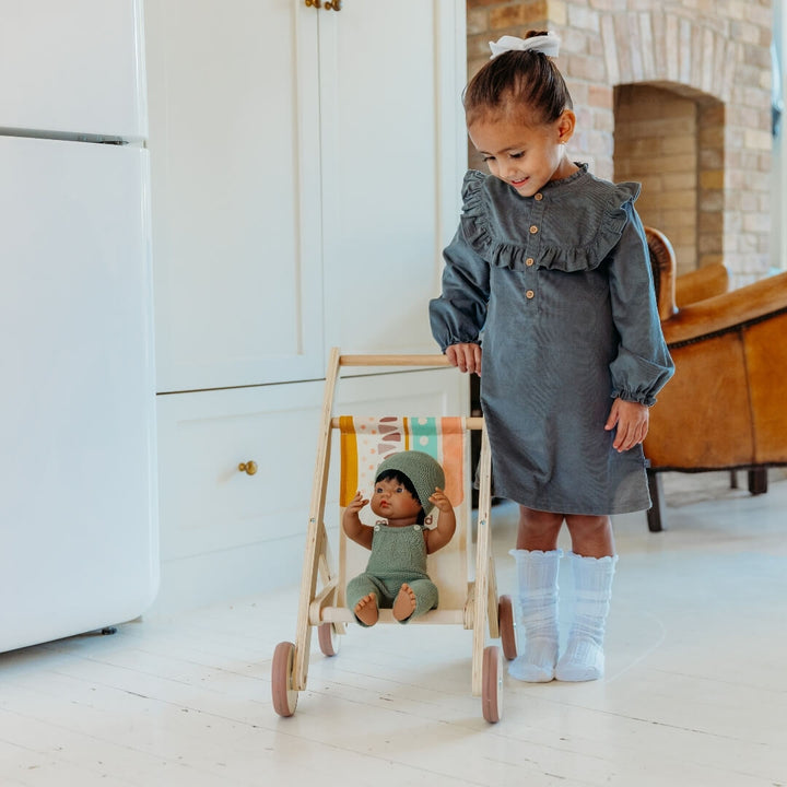 Child in a gray dress standing next to a doll in a wooden doll stroller by Miniland, made in Spain from sustainable wood with a colorful fabric seat.
