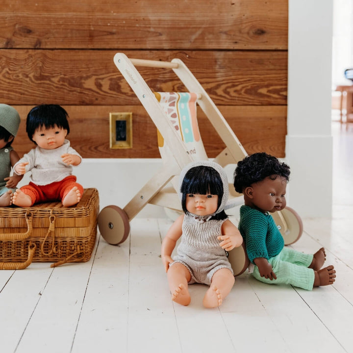 Three dolls with different hairstyles and outfits sitting on a wooden floor next to wooden doll stroller by Miniland, made in Spain from sustainable wood with a colorful fabric seat.