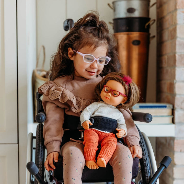 Child in a wheelchair holding a Miniland doll wearing glasses and knitted cream sweater and red pants set for 15-inch Miniland Dolls, made from recycled materials with a soft, handmade look—perfect for pretend play. 