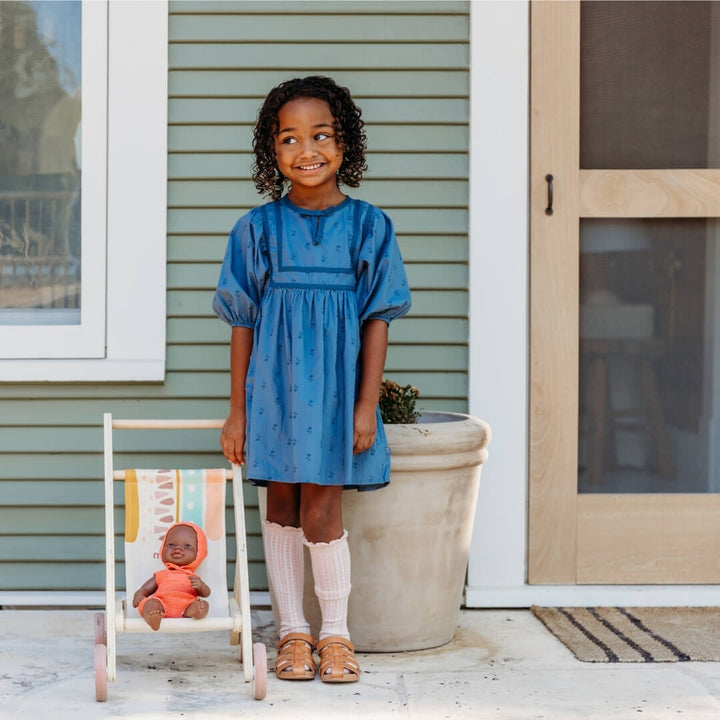 Young girl in a blue dress standing next to a Wooden doll stroller by Miniland, made in Spain from sustainable wood with a colorful fabric seat, outdoors.