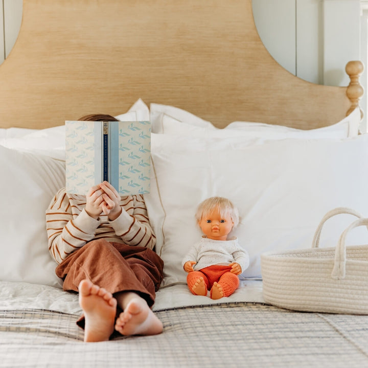 Child reading a book on a bed with a Miniland Realistic doll next to them who is wearing the knitted cream sweater and red pants set for 15-inch Miniland Dolls, made from recycled materials.
