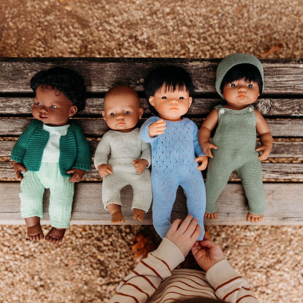 Four Miniland Realistic Montessori dolls in knitted outfits on a wooden surface with a hand reaching towards one of them.