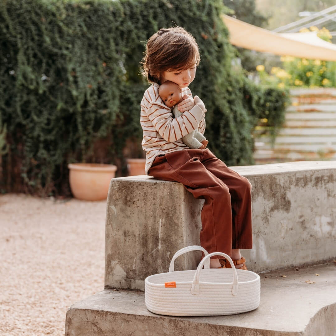 A child holds a Miniland baby doll. A Miniland doll carrier lays on the ground nearby.