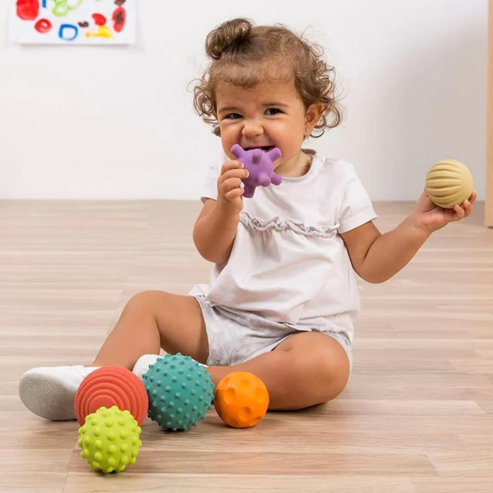 A toddler playing with a set of Miniland Natural Rubber Sensory Balls, holding 2 in their hands. The set includes six colorful textured balls in various shapes and sizes, designed for babies and toddlers to grasp, roll, and explore.
