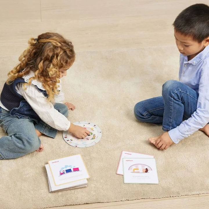Two children sit on a light-colored rug playing the Miniland Spin & Chill  mindfulness and relaxation game. One child spins a colorful activity wheel while the other watches. Several illustrated activity cards are spread out in front of them.