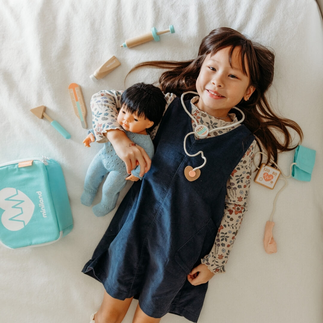A girl holding her Miniland doll laying on white blanket, surrounded by pieces from the Miniland wooden doctor kit.