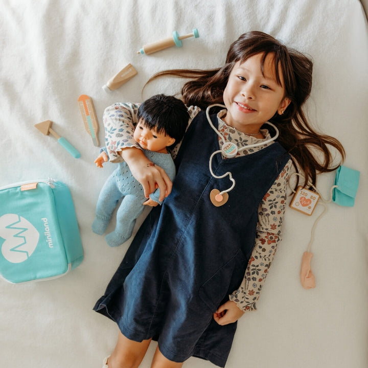 A girl holding her Miniland doll laying on white blanket, surrounded by pieces from the Miniland wooden doctor kit.