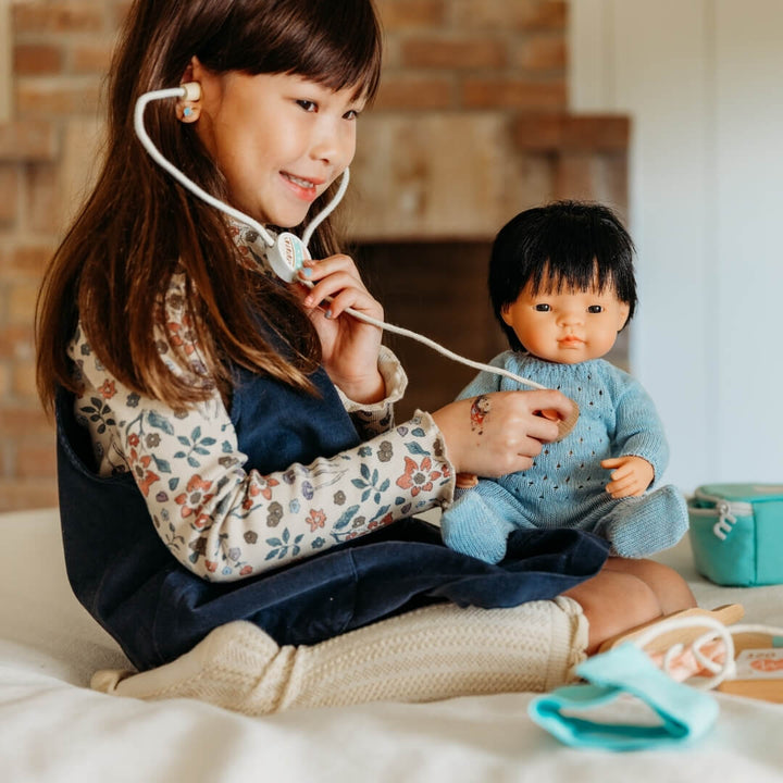 Child playing with a Miniland Realistic Boy doll  in Blue knit pajamas for 15-inch dolls, made from recycled materials with a handmade look—part of Miniland's eco-friendly doll clothing collection and toy stethoscope on a bed.