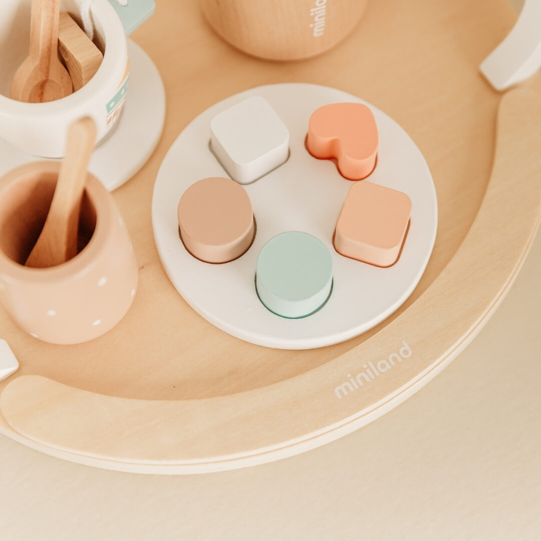 Children's toy set with wooden utensils and colorful plastic shapes on a beige tray, featuring the brand 'miniland'.