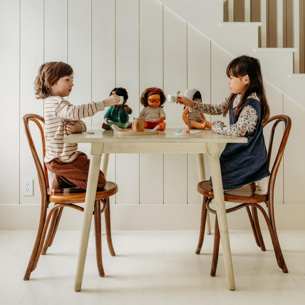 Two children sitting at a white wooden table, holding teacups from the Miniland tea set.