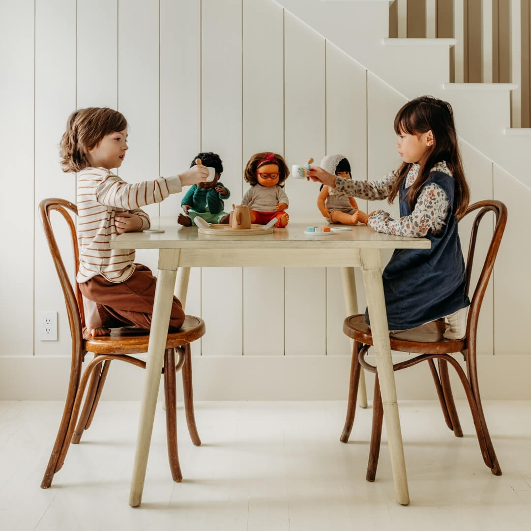 Two children sitting at a white wooden table, holding teacups from the Miniland tea set.