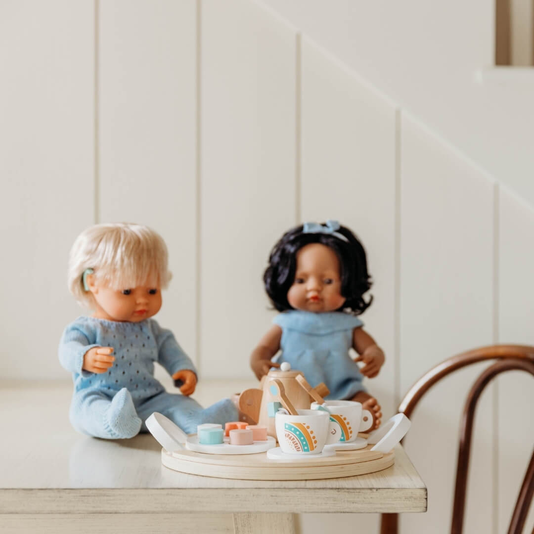 Two dolls in blue knit outfits sitting on a table next to the Miniland wooden tea set.