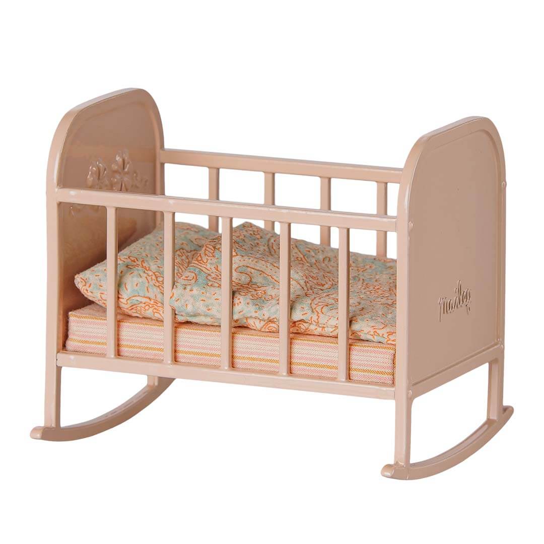 A small dollhouse rocking crib in a muted coral color, styled with a patterned blue and orange paisley quilt and striped mattress, on a solid white background.