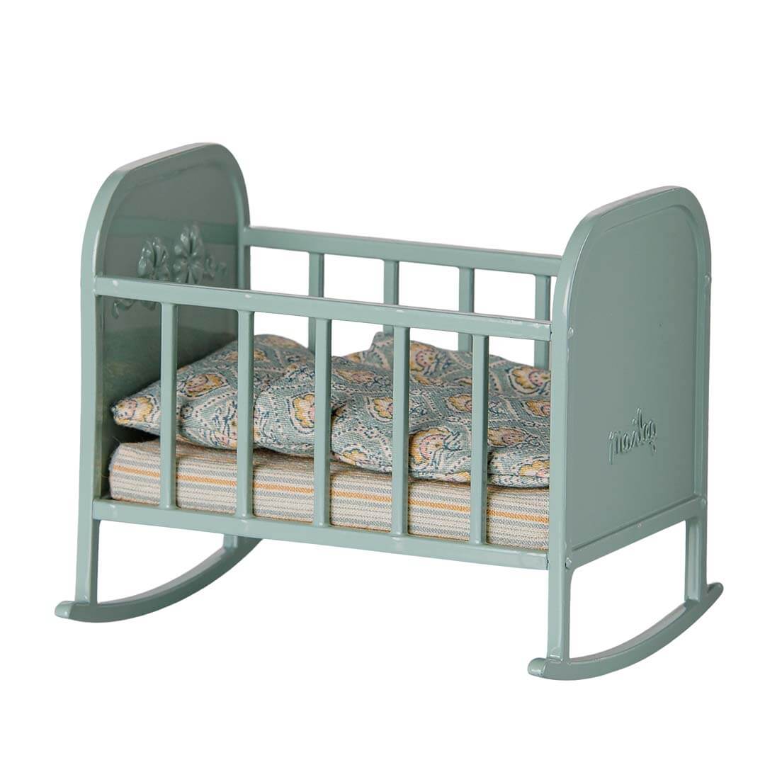 A small dollhouse rocking crib in a muted sage green color, styled with a patterned blue and orange paisley quilt and striped mattress, on a solid white background.