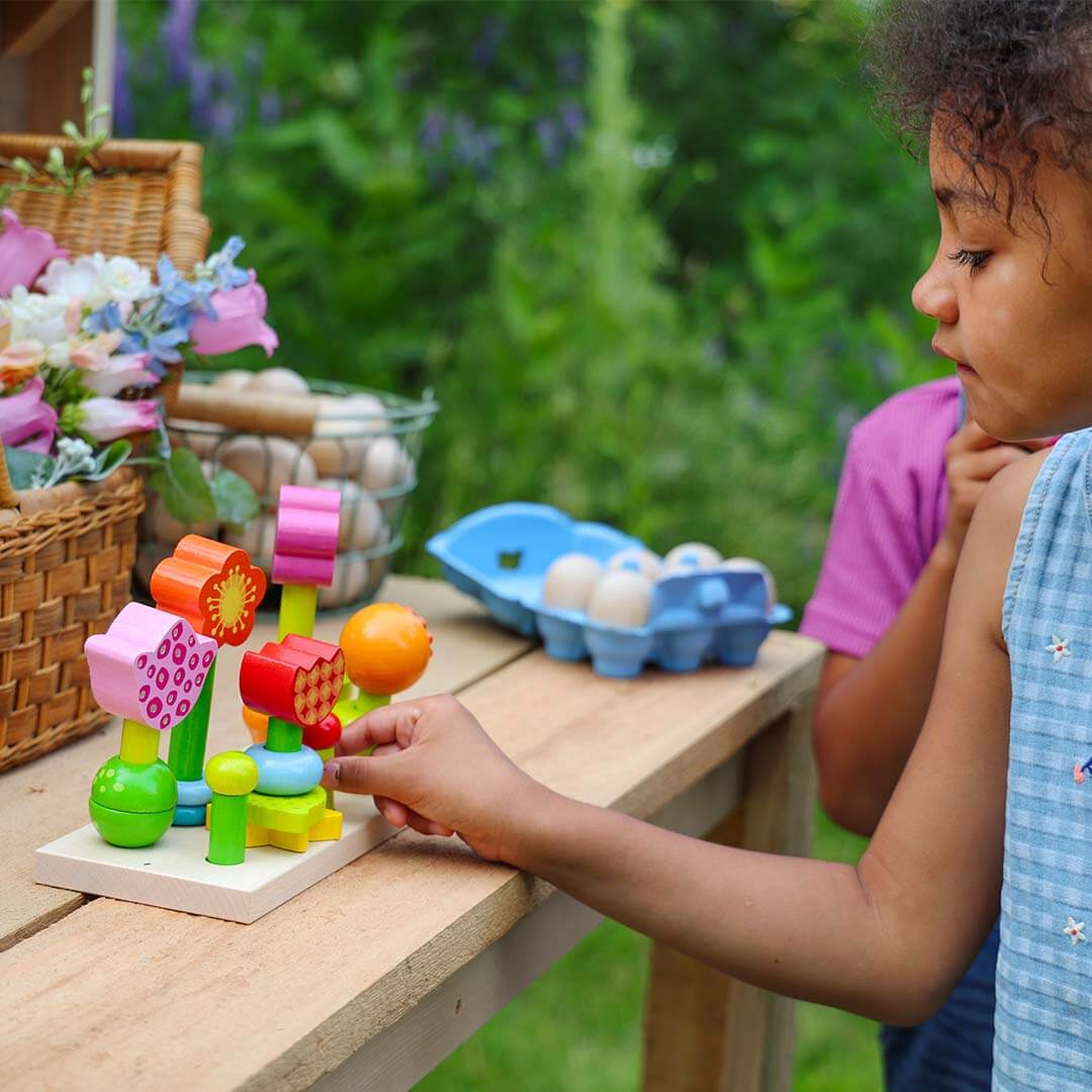 A child's hand plays with a colorful wooden flower sorting toy on a light wood table outdoors, with a basket of flowers, egg cartons, and another child partially visible in a garden setting.