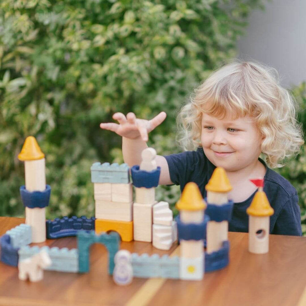 Child playing with PlanToys wooden castle block set arranged as a fortress with blue and yellow towers, arches, walls, and small wooden figures including a king, queen, knight, and horse. Bella Luna Toys