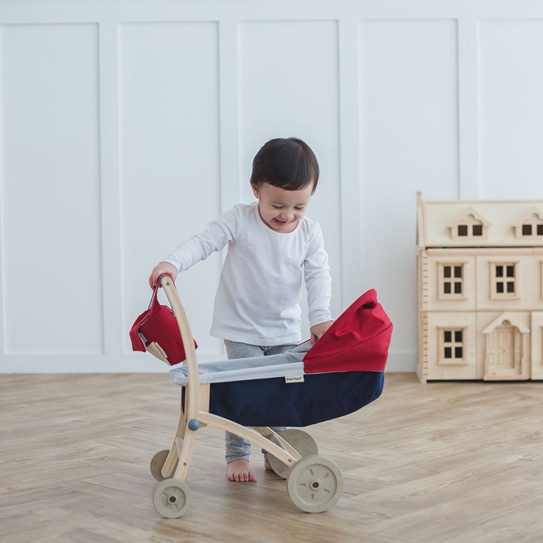 Smiling toddler peeking into the PlanToys Doll Stroller, interacting playfully with the wooden pushcart in a cozy playroom setting.