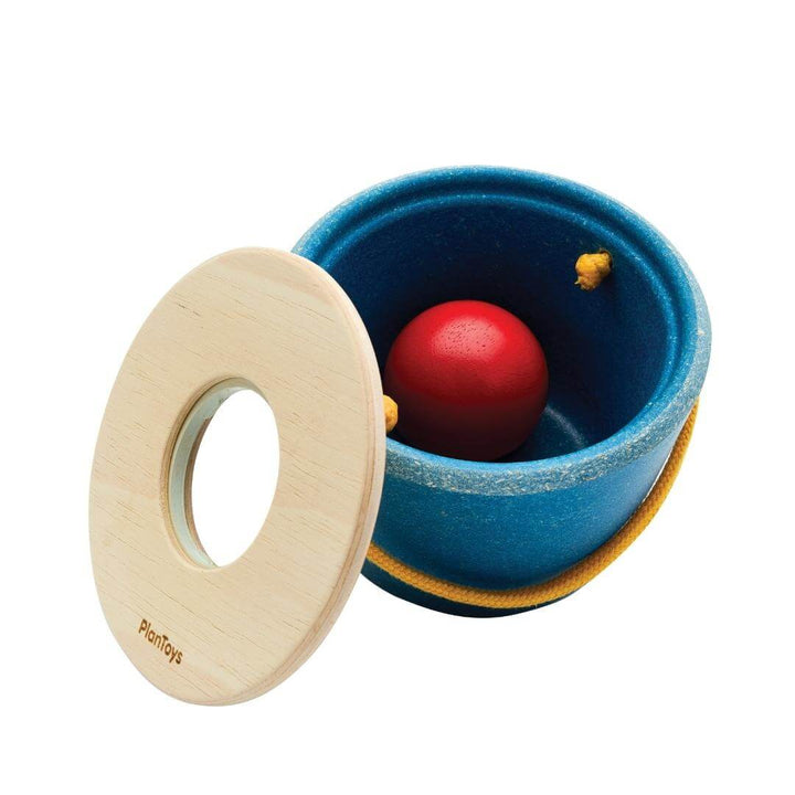 PlanToys Montessori Pop n Drop Bucket in blue with yellow handle, featuring a red wooden ball on top, designed for Montessori cause-and-effect play shown with the lid leaning on one side. 
