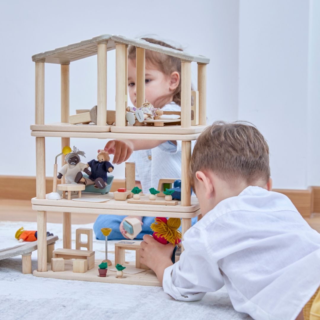 Two children playing with a multi-level wooden dollhouse, arranging furniture and figures in a modular PlanToys PlanSpace home setup.