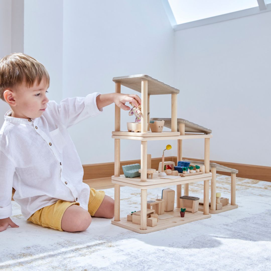 Young child playing with a multi-level wooden dollhouse, arranging furniture and figures in a modular PlanToys PlanSpace home setup.