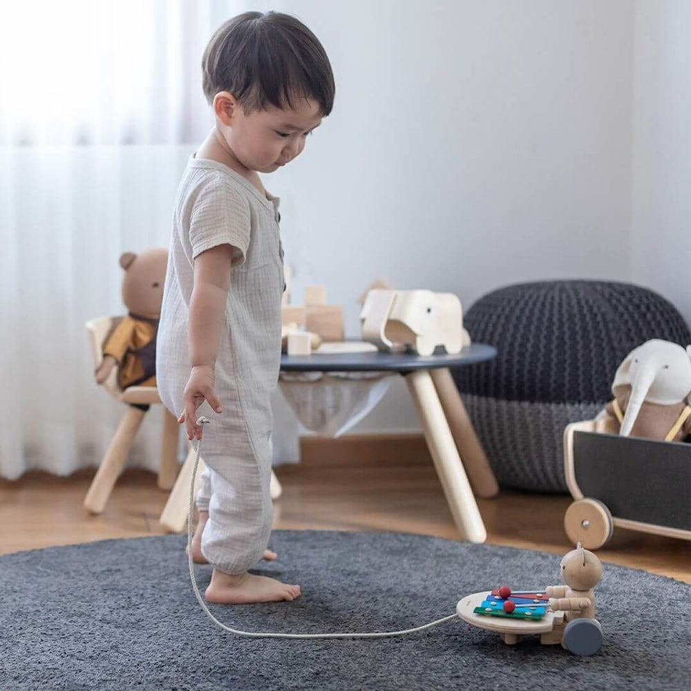 Child in a playroom setting looking at the wooden PlanToys pull-along bear tapping a colorful xylophone with red mallets, mounted on wheels with a pull cord attached. Bella Luna Toys.