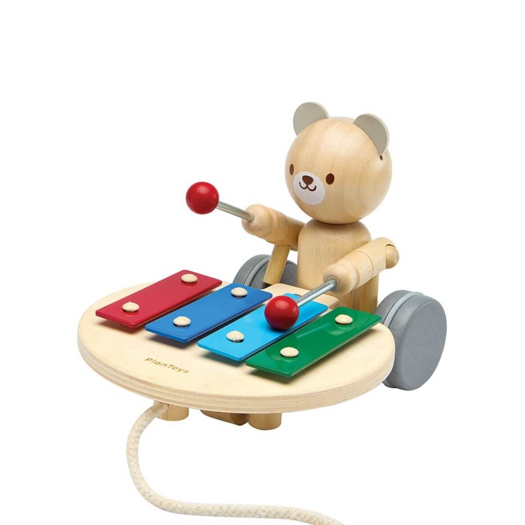 Wooden PlanToys pull-along bear tapping a colorful xylophone with red mallets, mounted on wheels with a pull cord attached. Bella Luna Toys.